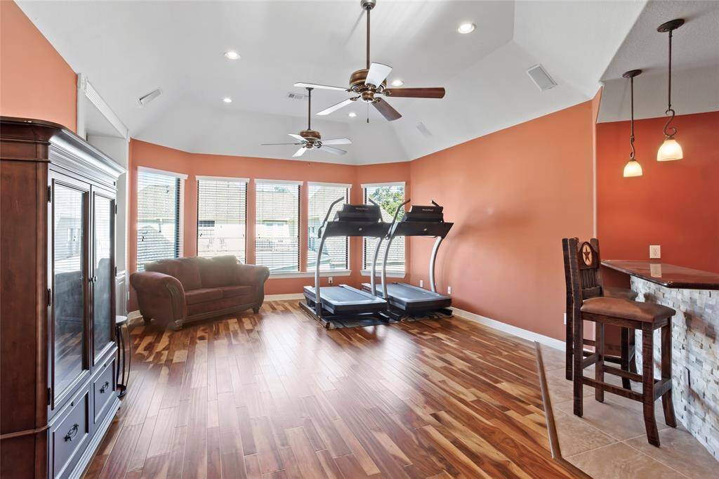 This spacious room features warm brazilian wood flooring, a cozy seating area, and large windows for natural light. It includes a full bar area with seating, perfect for relaxation or entertaining. The ceiling fans and recessed lighting add comfort and ambiance.