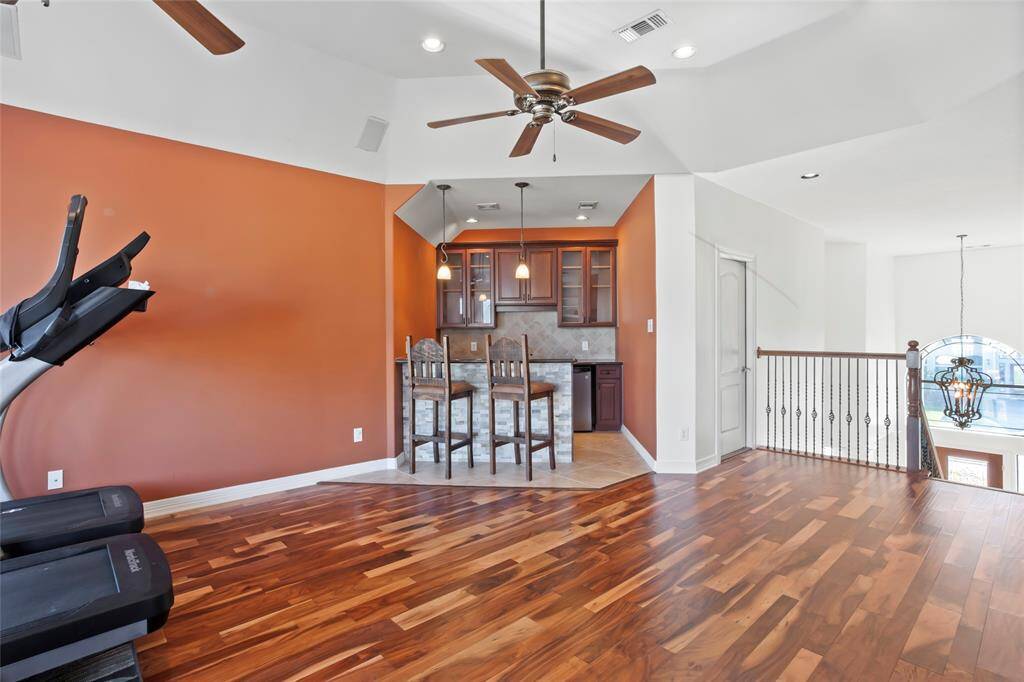 This spacious room features rich brazilian wood flooring and a burnt orange accent wall, perfect for a home gym or entertainment area. It includes a cozy bar nook with pendant lighting, a ceiling fan, and an open view to the lower level.