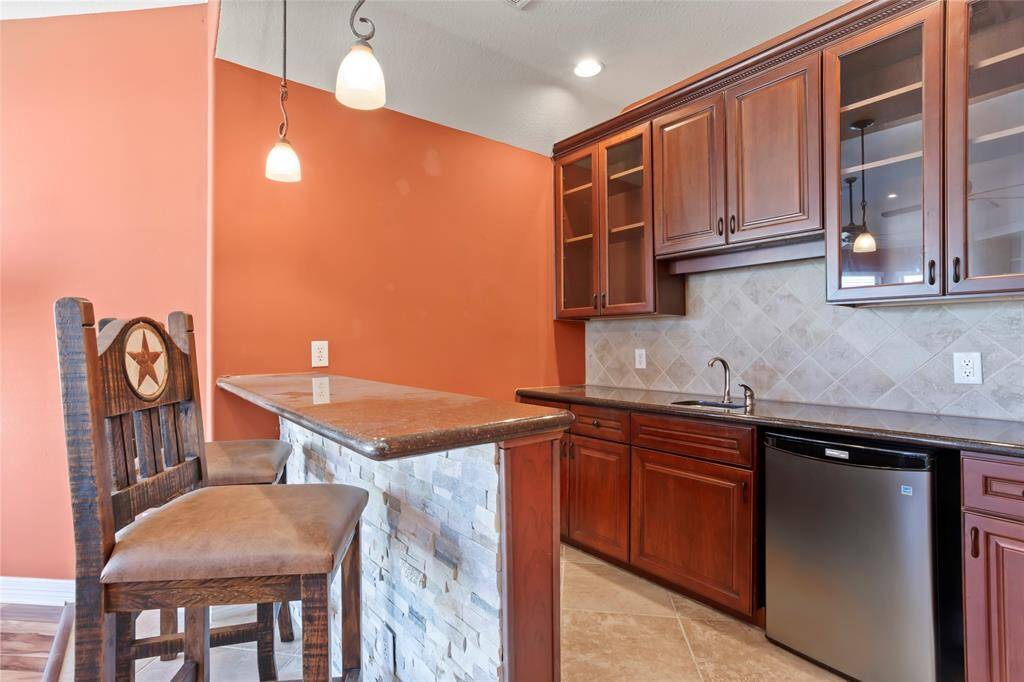 A cozy full bar area with burnt orange walls and elegant dark wood cabinetry. It features a small sink, a mini-fridge, dishwaster and a stylish stone countertop with pendant lighting. Perfect area for entertaining.