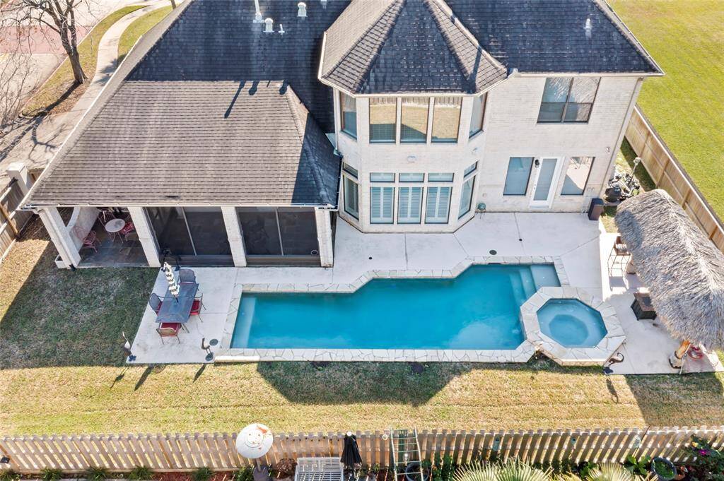 This aerial view showcases a spacious two-story home with a backyard featuring a rectangular swimming pool and adjacent hot tub. The patio includes a covered area, perfect for outdoor dining and relaxation, and a thatched-roof section adds a tropical touch. The yard is fenced, providing privacy and security.