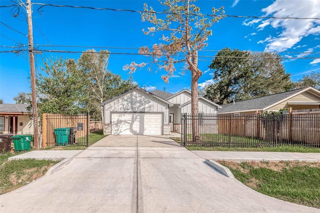 This photo shows a modern single-story house with a clean, white exterior and a two-car garage. It's set back from the street with a long driveway and is fenced on both sides. The property is surrounded by trees, providing a sense of privacy and nature.