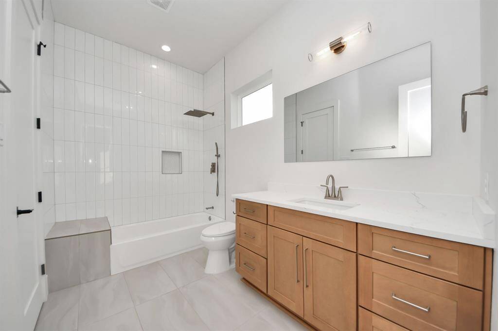The shower boasts grand floor-to-ceiling tiles, complete with a custom nook and bench for extra product storage. The vanity offers ample space, enhanced by upgraded wood-style cabinets that bring both elegance and functionality to the room.