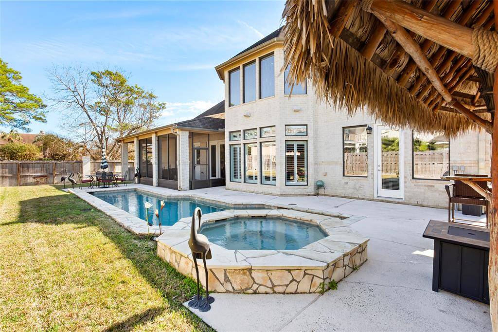 This photo showcases a spacious backyard featuring a swimming pool and a spa, surrounded by a patio area. The home has large bay windows and a screened porch, providing a great view of the outdoor space. A thatched-roof structure adds a tropical touch, making it ideal for relaxation and entertaining.