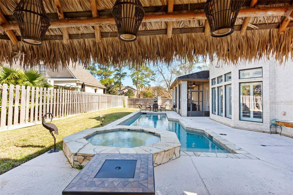 This photo showcases a backyard oasis featuring a modern pool and spa, surrounded by a spacious patio and a thatched-roof pergola. The outdoor area is perfect for relaxation and entertaining, with ample space and privacy provided by a wooden fence and lush greenery.