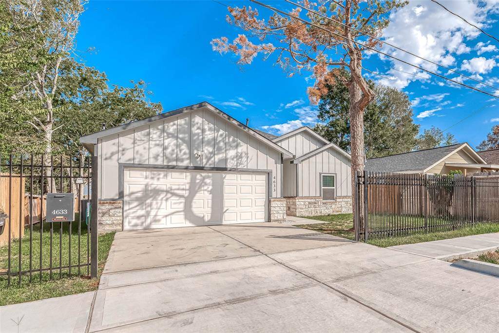 This photo shows a modern single-story home with a two-car garage and a fenced front yard. The exterior features a combination of light gray siding and stone accents. The driveway is spacious, and there are mature trees around the property, providing a welcoming and serene atmosphere.