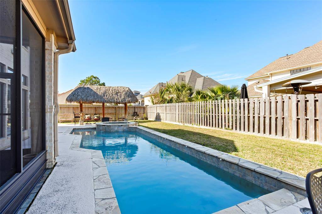 This backyard features a sleek rectangular pool with a stone border, a spa, a new wooden privacy fence, and a thatched-roof pergola, perfect for relaxation and outdoor gatherings.