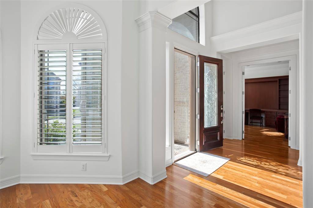 Bright entryway with hardwood floors, featuring a decorative glass front door and large windows with shutters. The space leads into an office with built-in shelving on the other side is a large sitting room offering a warm and inviting atmosphere for entertainment or relaxation.