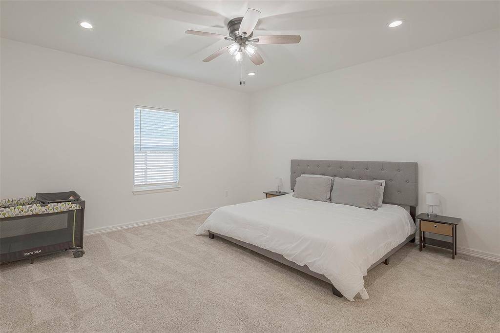 This spacious bedroom features neutral tones with a plush carpet, and ample natural light from a single window. It includes a ceiling fan, recessed lighting creating a cozy and inviting atmosphere.