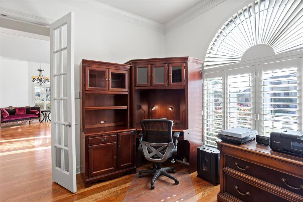 A bright home office with elegant hardwood floors and a large arched window with shutters. It features a classic wooden desk, ample storage, and flows into a spacious sitting area.