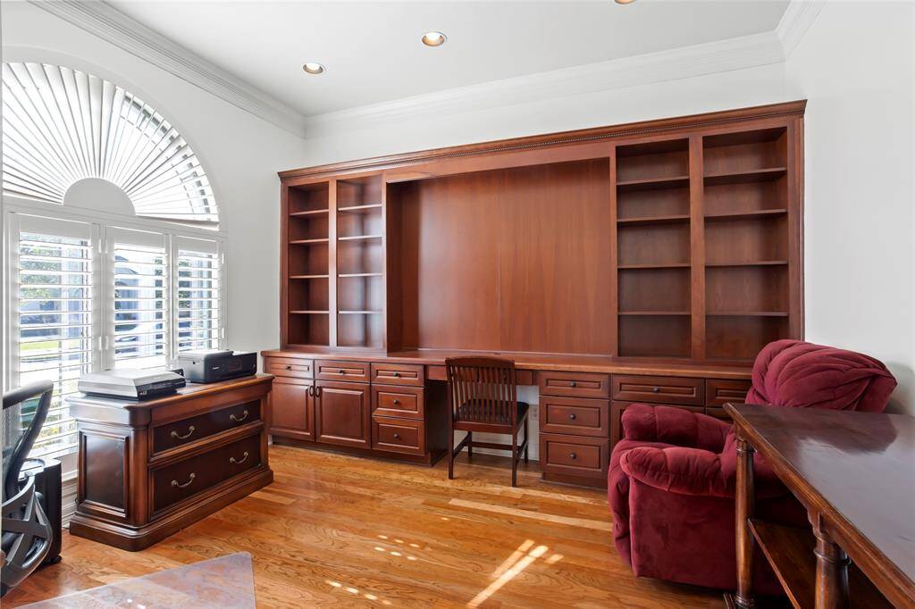 Bright home office with hardwood floors, built-in wooden shelves, and a large arched window with shutters.
