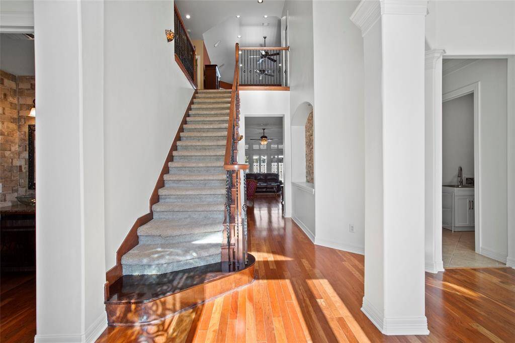 This elegant home entryway features a grand staircase with carpeted steps and wooden accents. The space is bright with high ceilings, polished hardwood floors, and is flanked by white walls and columns, leading into a spacious living area.