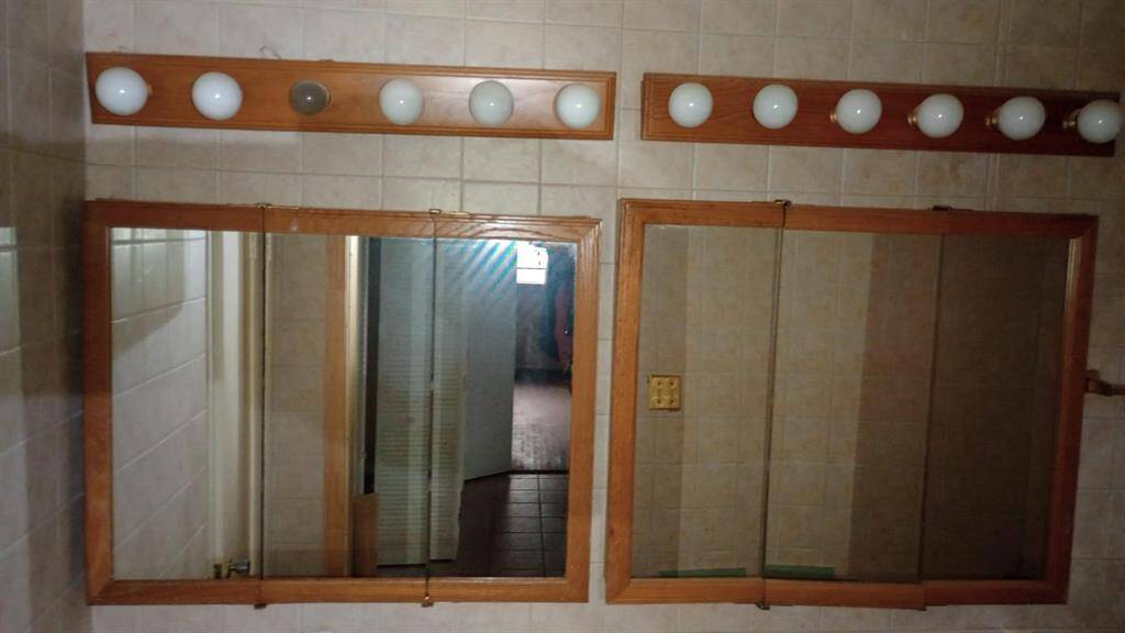 This bathroom features dual mirrors with wooden frames and ample lighting from rows of globe bulbs above. The surrounding walls are tiled in a neutral tone, creating a classic and functional space.