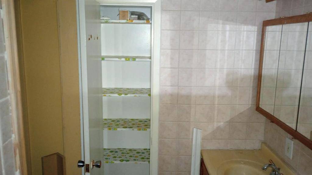 This photo shows a bathroom with a tiled wall and a beige countertop. There's a built-in shelf with patterned lining for storage, and a mirror above the sink. The space appears functional with some room for updates or personalization.