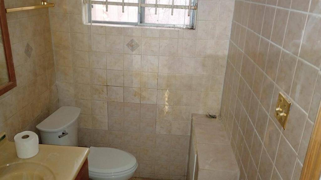 This is a compact bathroom with tiled walls and floor. It features a toilet, a small vanity with a sink, and a window for natural light. The tiles have a neutral color palette, and there is a wall-mounted mirror and towel rack.