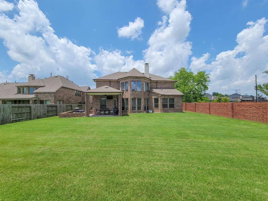Spacious backyard with a large, two-story brick house. There's a covered patio area perfect for outdoor dining or relaxation, and a vast lawn ideal for recreation or gardening. The property is enclosed by a combination of wooden and brick fencing, providing privacy and security.