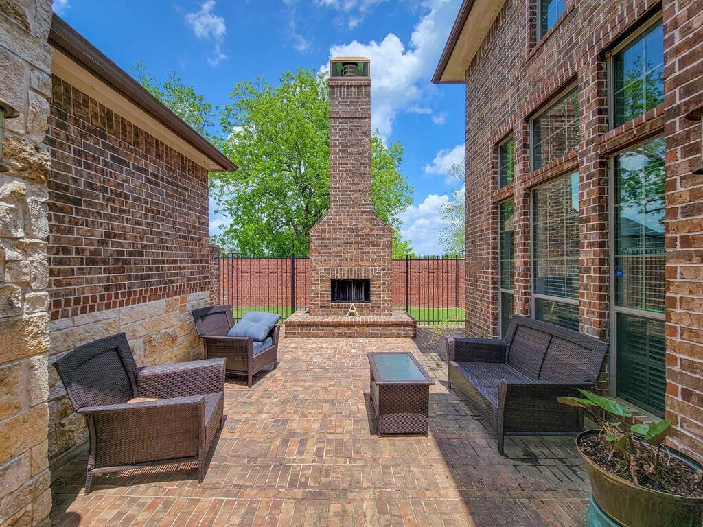 Outdoor patio with brick flooring and walls, featuring a central fireplace. It is furnished with comfortable wicker seating and a glass-top table, ideal for relaxing or entertaining. A brick privacy fence and lush greenery provide an inviting, secluded atmosphere.