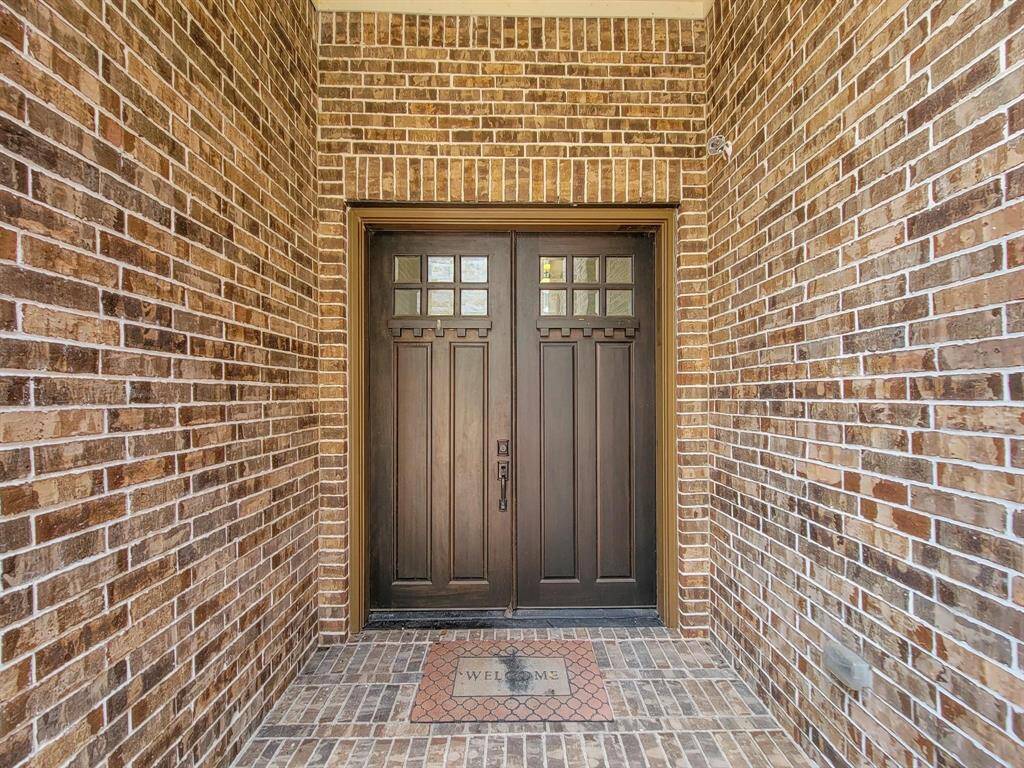 A welcoming front entrance features a sturdy wooden double door with decorative glass panels, framed by classic red brick walls and complemented by a "Welcome" mat on the brick-patterned flooring.