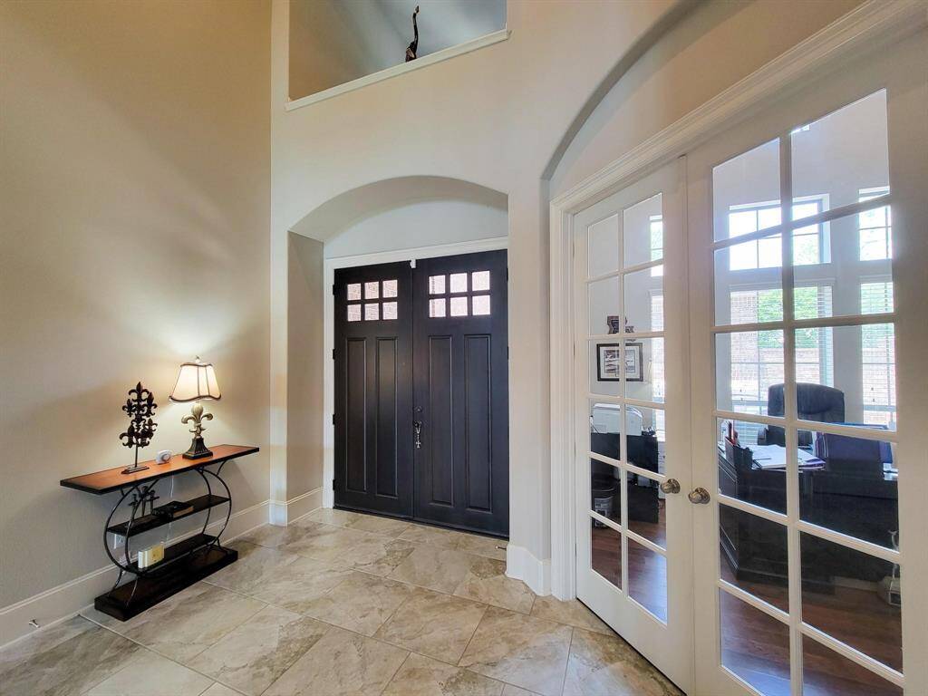 Bright, welcoming entryway featuring elegant double doors, a stylish side table with a lamp, and glass-paneled doors leading to a well-lit office space. The high ceiling and neutral tones create an open and airy feel.