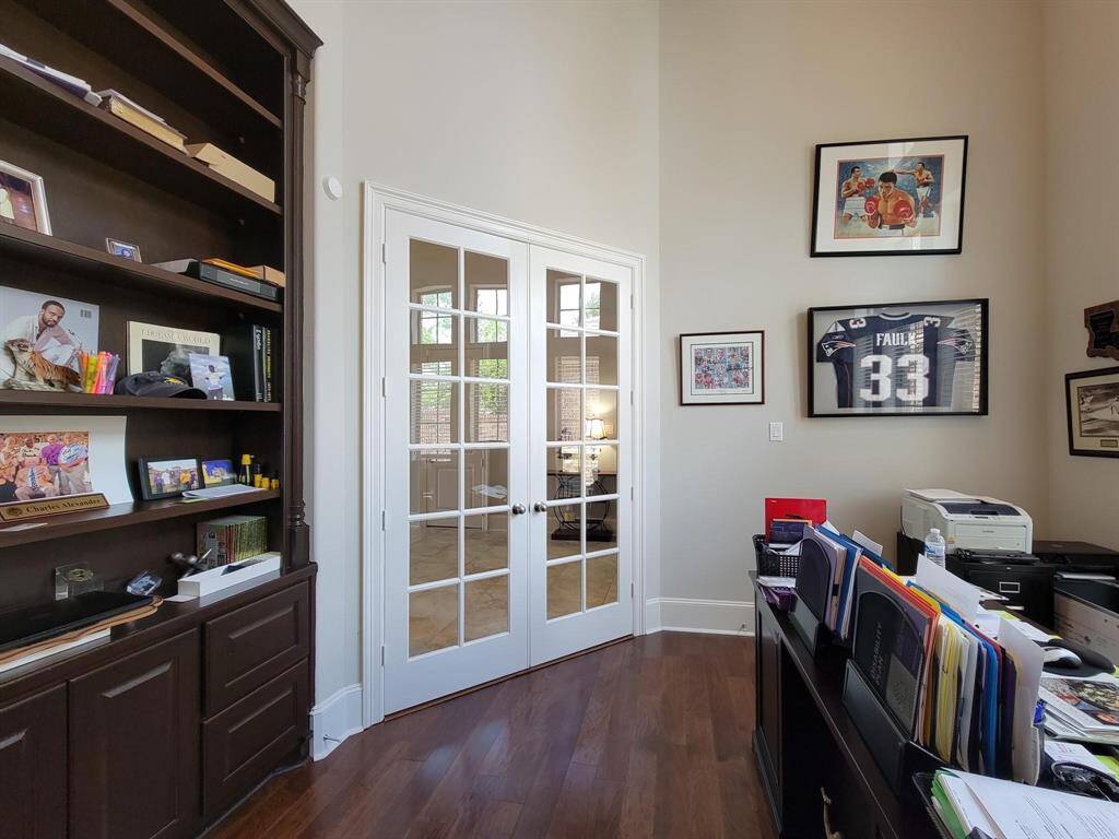 Cozy home office with built-in dark wood shelves, French doors, and framed sports memorabilia. The space is well-lit, has hardwood floors, and offers a view into an adjacent room.