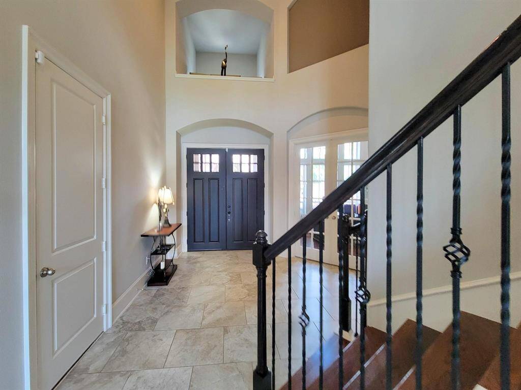 Spacious entryway with high ceilings, elegant tile flooring, and a dark wooden door. A stylish staircase with wrought iron balusters adds sophistication, while ample natural light enhances the welcoming atmosphere.