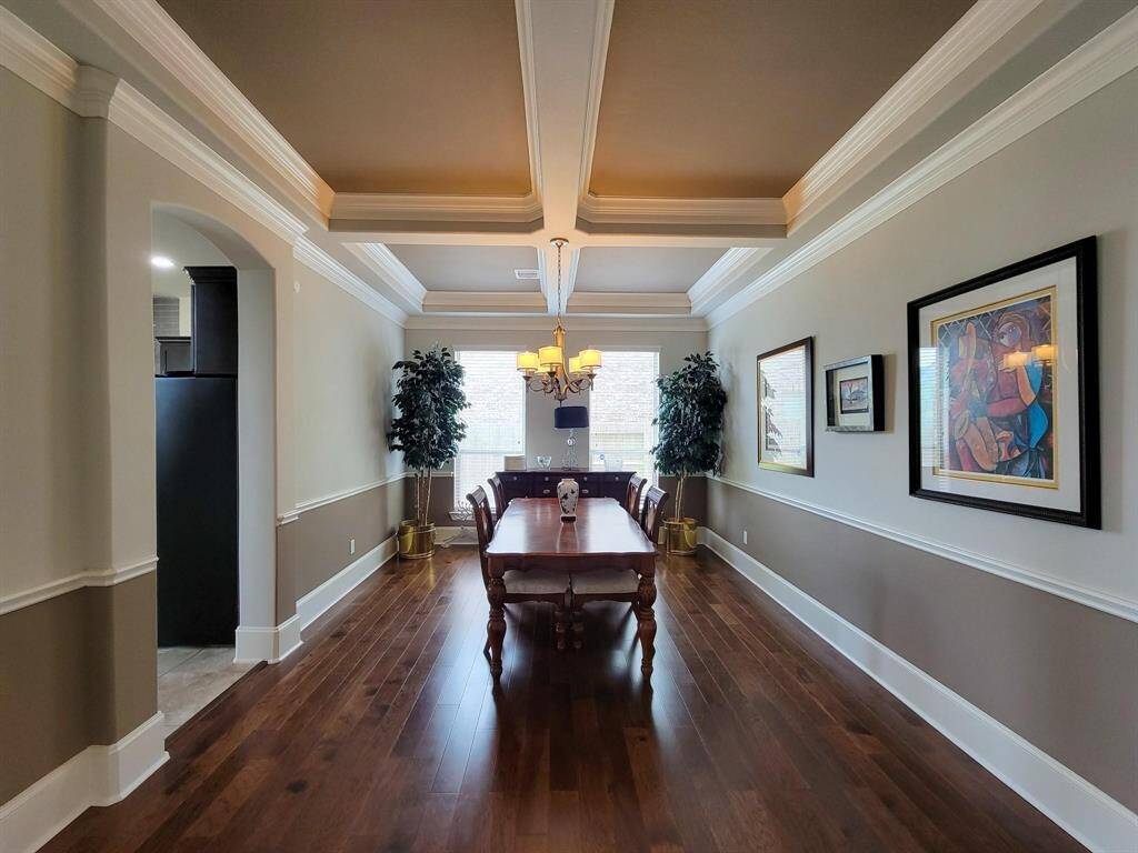 This elegant dining room features a coffered ceiling, rich hardwood floors, and ample natural light. It includes a central wooden dining table, decorative moldings, and tasteful artwork on the walls, creating a warm and inviting atmosphere.