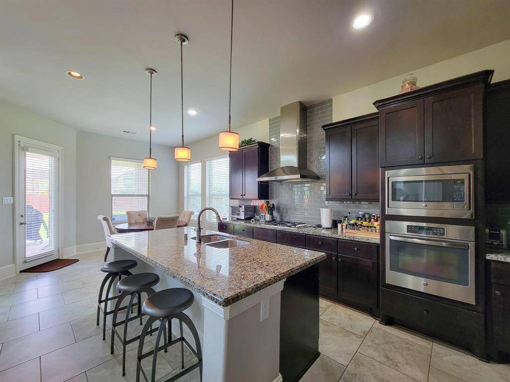 Spacious modern kitchen with granite countertops, dark wood cabinets, and stainless steel appliances. Features a large island with seating, pendant lighting, and a cozy breakfast nook. Ample natural light from multiple windows and a door leading outside.