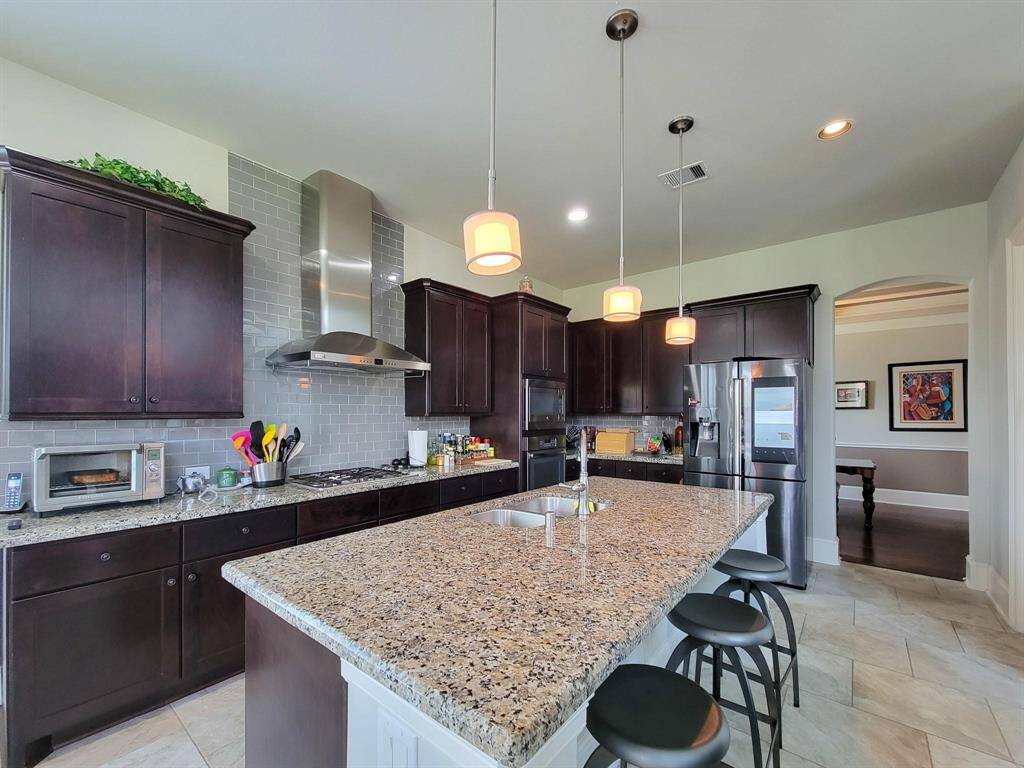 This kitchen features a spacious island with granite countertops, dark wood cabinetry, and stainless steel appliances. It has modern lighting fixtures and an open layout, ideal for entertaining and meal preparation.