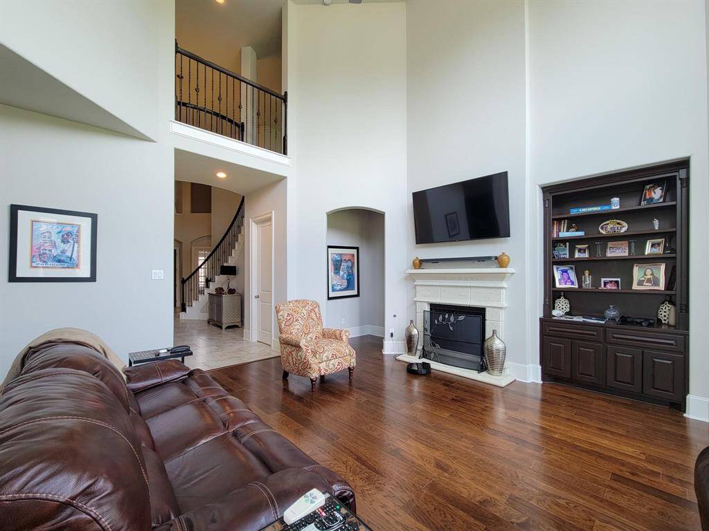 This spacious living room features high ceilings, a sleek hardwood floor, and a cozy fireplace. It includes built-in shelving, a large TV, and elegant staircase access, offering a comfortable and stylish setting.
