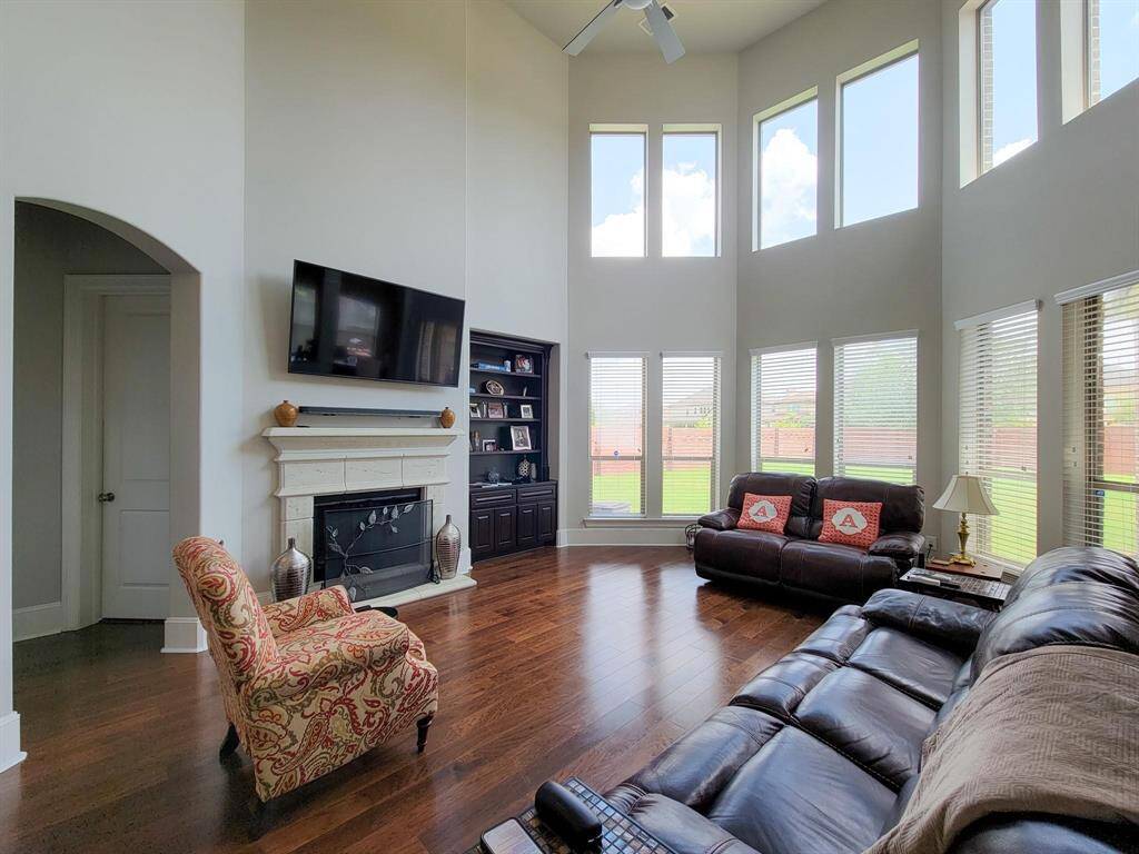 This spacious living room features high ceilings, large windows for natural light, and elegant hardwood floors. It includes a cozy fireplace, mounted TV, and ample seating, making it perfect for family gatherings.