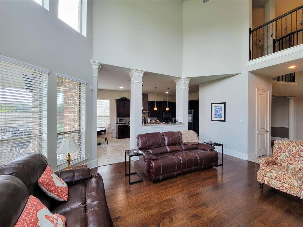 This spacious living area features high ceilings, large windows for natural light, and elegant wood flooring. It opens to a modern kitchen with dark cabinetry and a breakfast bar, creating an inviting, open-concept space ideal for entertaining.