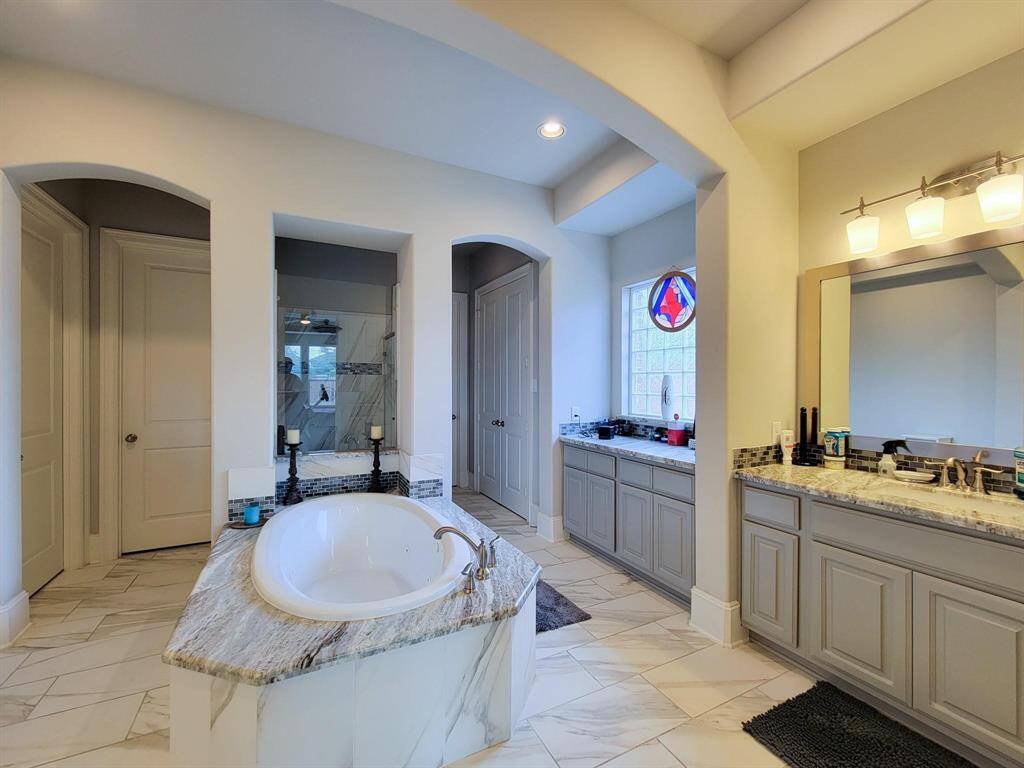 This spacious bathroom features a luxurious freestanding tub, dual vanities with granite countertops, and elegant tile flooring. The room is well-lit with recessed lighting and a large window, offering a relaxing and modern atmosphere.