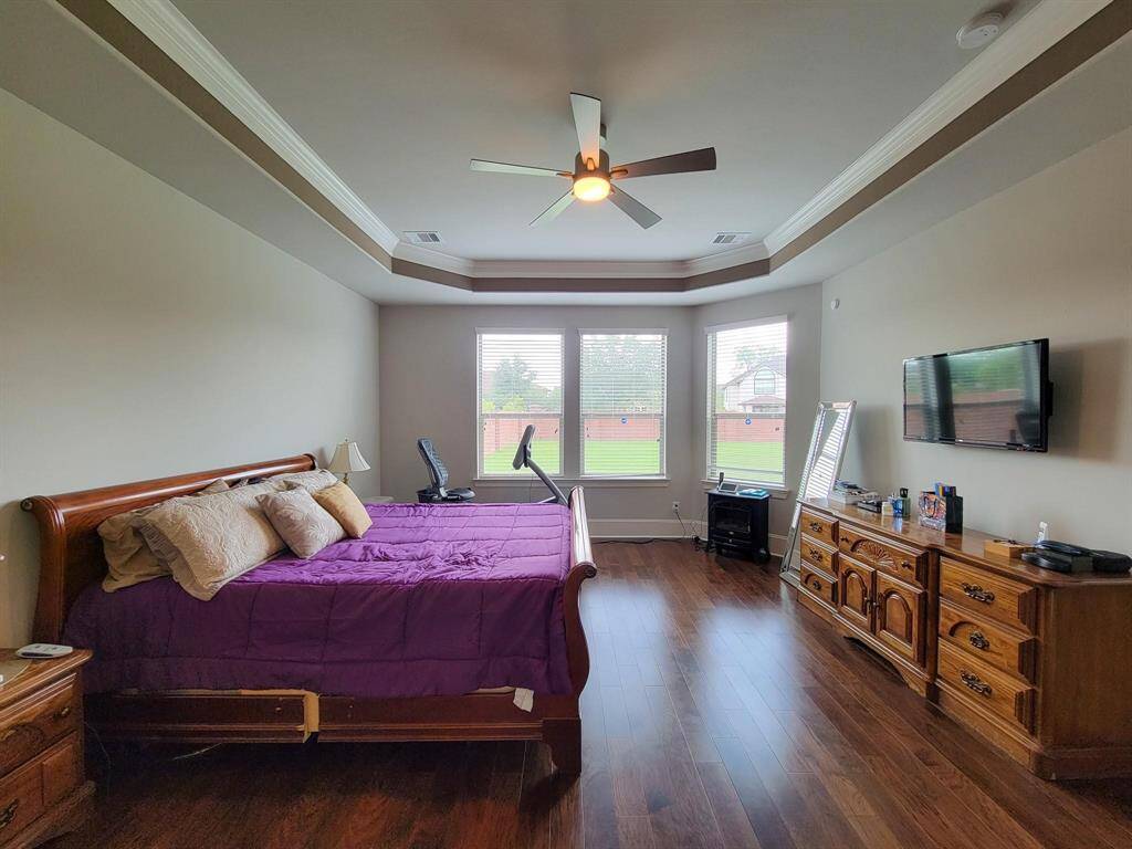 Spacious bedroom featuring a tray ceiling, large windows with garden views, and rich wood flooring. The room includes a mounted TV, a cozy bed with a wooden frame, and ample storage with matching wooden furniture.