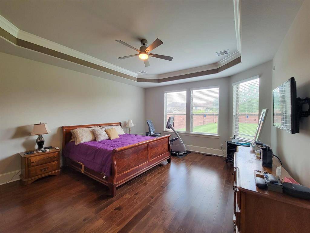 Spacious bedroom with hardwood floors, featuring a tray ceiling and large windows providing ample natural light. It includes a ceiling fan, a wooden bed frame, and offers a view of the backyard.