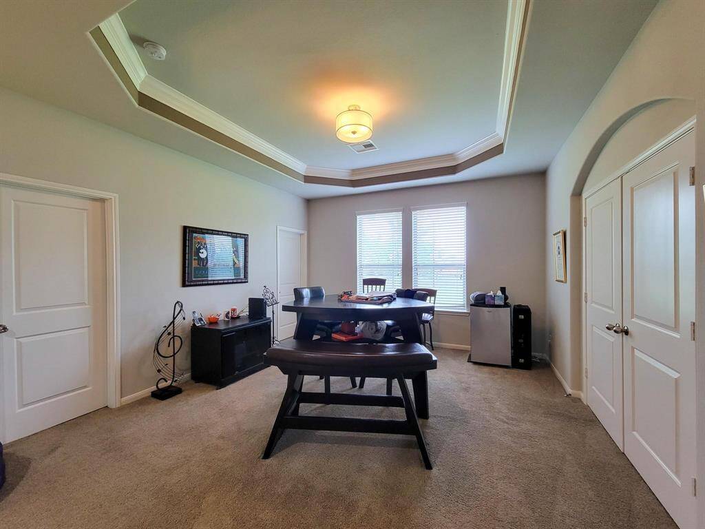 This room features a spacious layout with a tray ceiling and abundant natural light from large windows. It includes double doors, carpeted flooring, and neutral walls, ideal for use as an office or study.