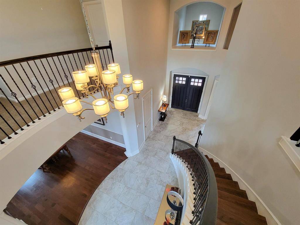 This elegant foyer features a grand curved staircase with dark wood steps and wrought iron railing, leading to a balcony overlooking the entrance. The space is illuminated by a large chandelier, and the double front doors open to a tiled floor, creating a welcoming and sophisticated ambiance.
