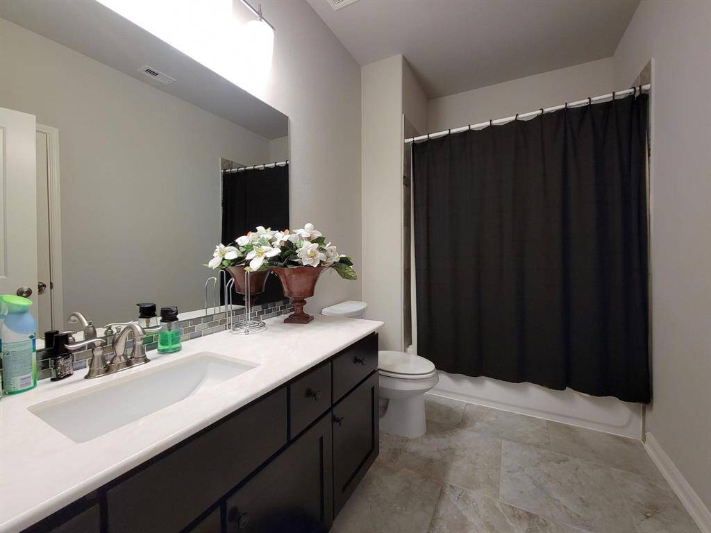 This bathroom features a sleek design with a modern vanity, a large mirror, and a black shower curtain. The countertop is spacious with decorative accents, and the room has neutral tile flooring.