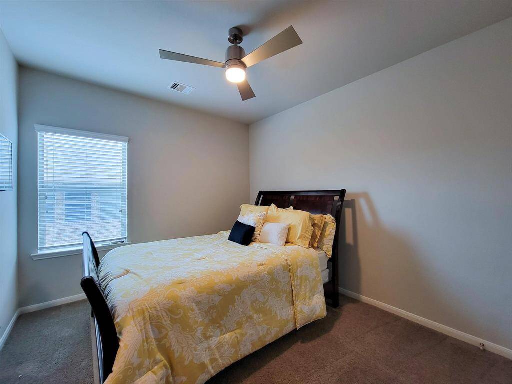 This cozy bedroom features neutral walls, plush carpeting, and a ceiling fan. Natural light streams in through a window with blinds, complementing the comfortable bed with stylish bedding.