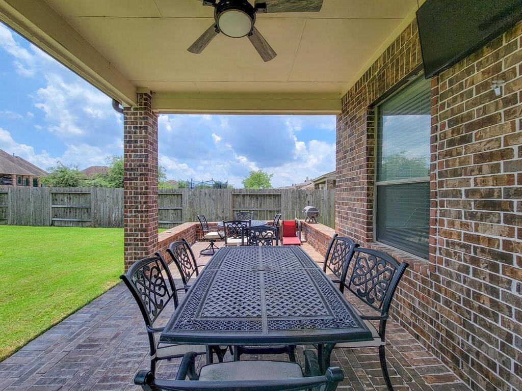Covered brick patio with elegant outdoor dining furniture, a ceiling fan, and a view of a well-maintained fenced backyard, ideal for entertaining and relaxation.
