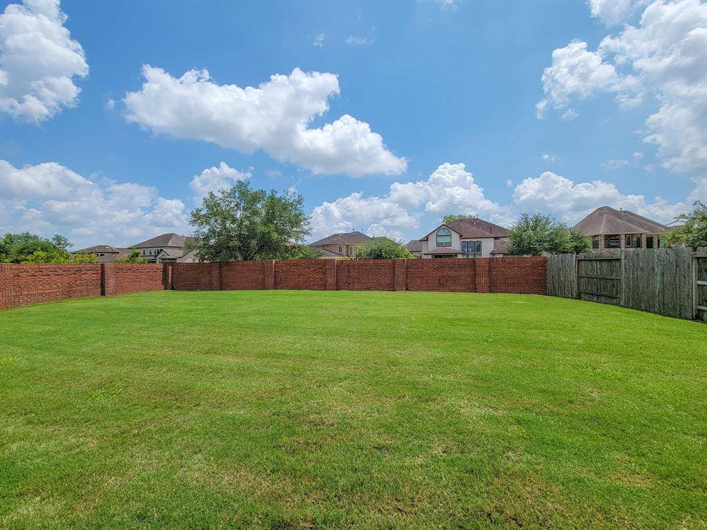Huge, spacious backyard with lush green lawn, surrounded by a mix of wooden and brick fencing, offering privacy and a clear view of neighboring houses under a bright, blue sky.