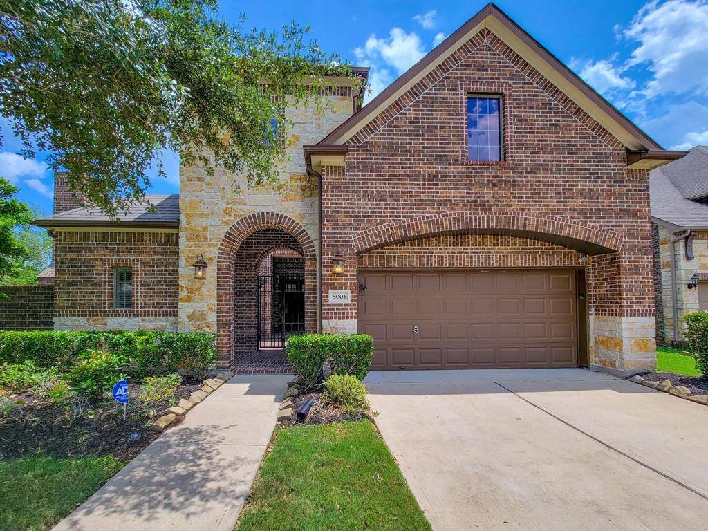 This is a charming brick and stone two-story home with a welcoming arched entryway and a two-car garage. It features a well-maintained front yard with lush landscaping, creating an inviting curb appeal.