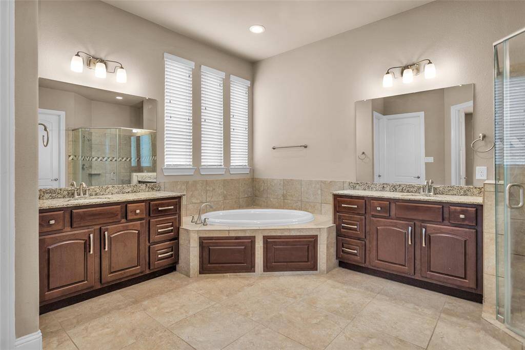 This luxurious, yet spacious bathroom features dual vanities with granite countertops, a central soaking tub, and a glass-enclosed shower. Natural light streams through the windows, highlighting the elegant tile flooring and warm wood cabinetry.