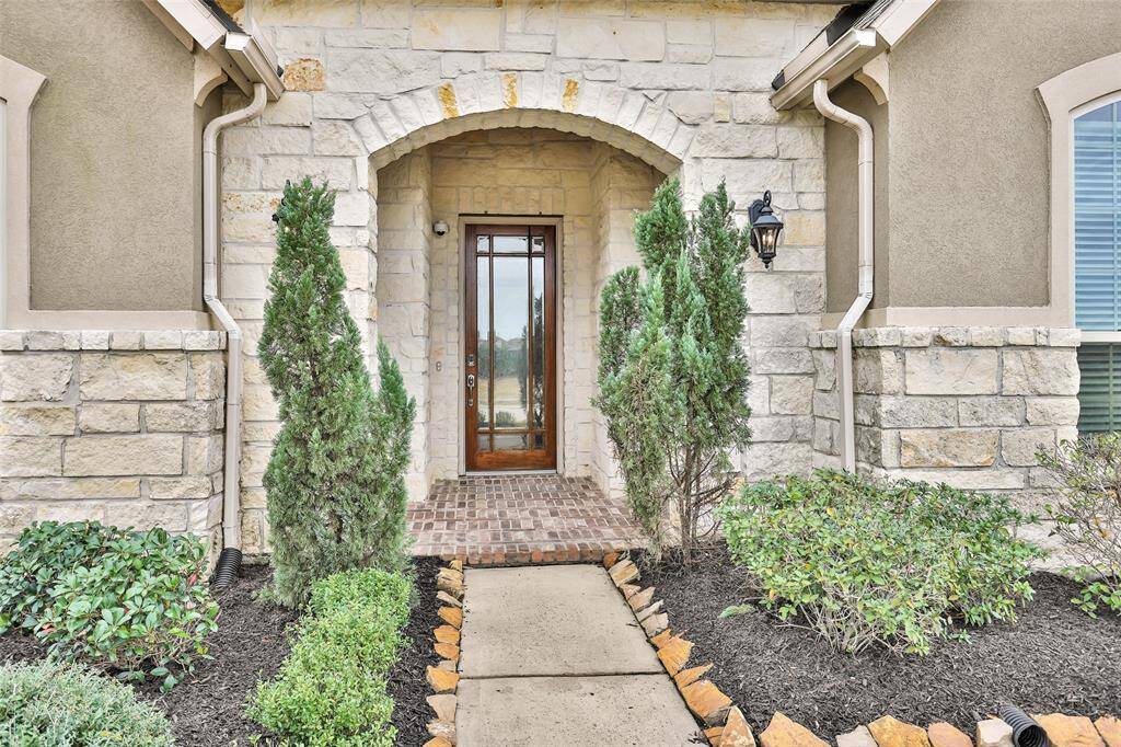 This inviting entryway features a charming stone archway, elegant wooden door with glass panels, and neat landscaping with shrubs and trees, creating a warm welcome.