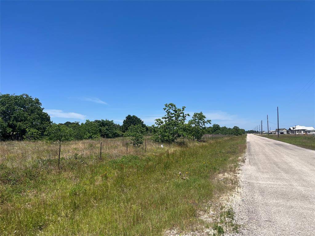 This photo shows a rural setting with a grassy plot of land bordered by a fence, alongside a private gravel road. The area is open and spacious with scattered trees, offering a quiet, natural environment.