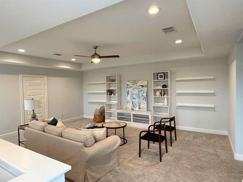This spacious living area features neutral carpeting, built-in shelving, and recessed lighting. It includes a ceiling fan, comfortable seating, and modern decor, making it ideal for relaxation or entertaining.