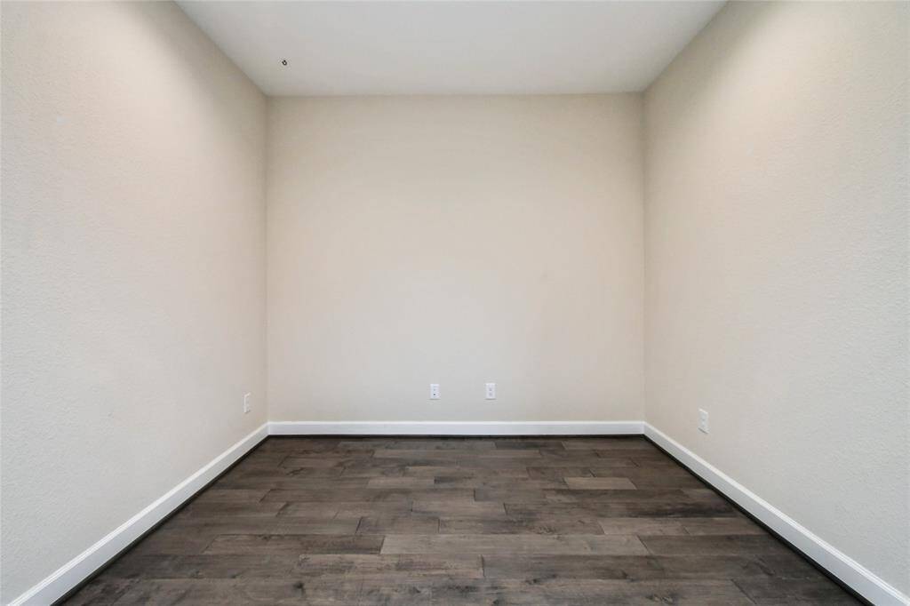 This photo shows a small, unfurnished room with light beige walls and dark wood flooring. It features a simple design with baseboards and electrical outlets, ideal for a home office or storage space.