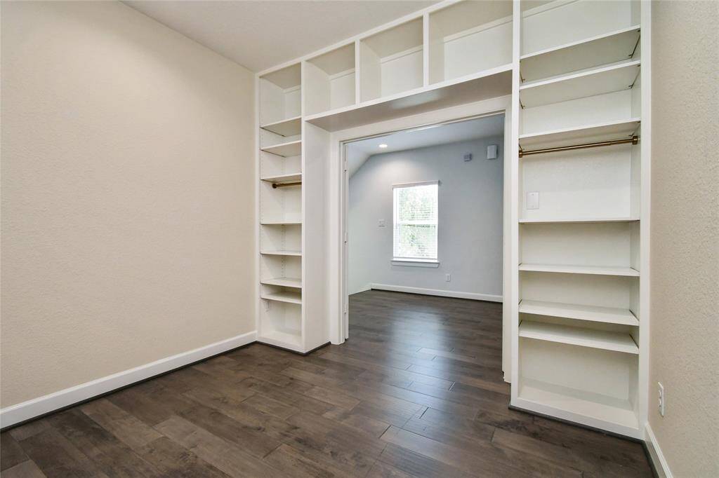 This photo shows a spacious room with dark hardwood floors and built-in white shelving units. The room is well-lit with a window letting in natural light, and there is a cozy nook area, making it perfect for a home office or reading space.