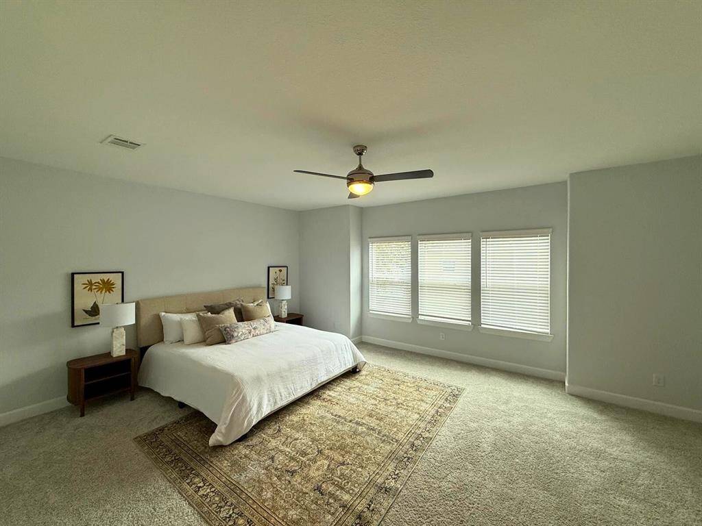 Spacious, light-filled bedroom with neutral tones, featuring a ceiling fan and three large windows. Includes a cozy carpet and a stylish area rug, complemented by modern bedside tables and lamps.