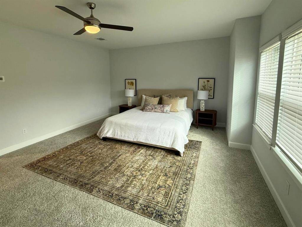 Spacious bedroom with neutral tones, featuring a large bed, two bedside tables, and a ceiling fan. The room is carpeted and has ample natural light from the windows.