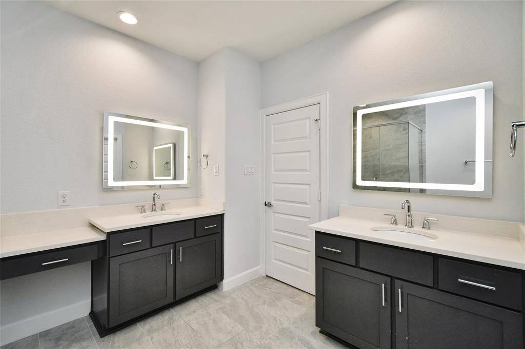 This bathroom features dual vanities with modern dark cabinetry and illuminated mirrors. The space is bright with a neutral color palette, and there's a separate shower area visible.