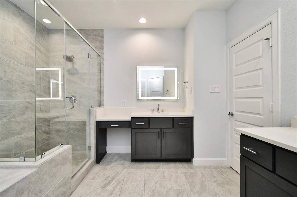 This modern bathroom features a spacious glass-enclosed shower with sleek tile work. It has a large vanity with dark cabinets and a lighted mirror, complemented by light-colored walls and flooring. The overall look is clean and contemporary.